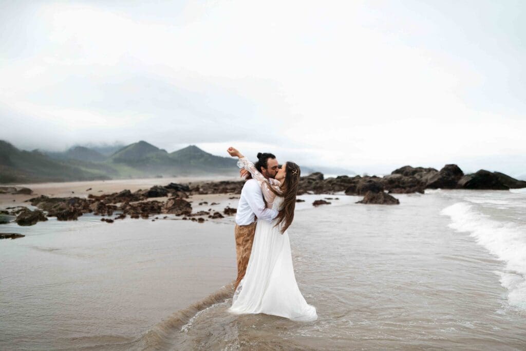 a couple kiss on the beach with hills and rocks behind them during their elopement day