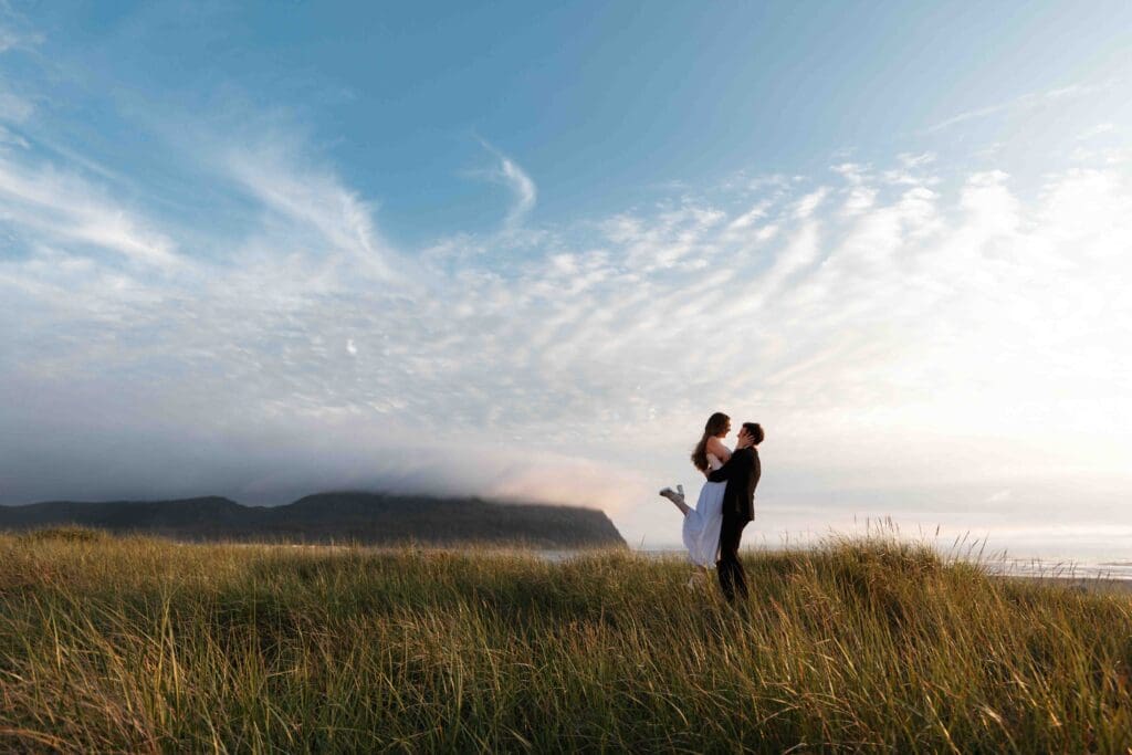 a groom lifts up his bride in the tall beach grass on the coast